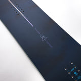 Nidecker Ora Women's Snowboard 2025 - A dark blue snowboard with a white stripe down the middle and a geometric logo in purple at the top.