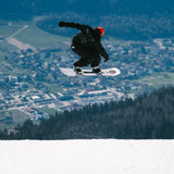 Nidecker Thruster Snowboard 2025 - A white snowboard with black bindings and a black strap on top of a snowy mountain.