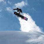 Nidecker Venus Plus Women's Snowboard 2025 - A black snowboard with a pink and blue logo on top, flying through the air at an angle.
