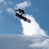 Nidecker Venus Plus Women's Snowboard 2025 - A black snowboard with a pink and blue logo on top, flying through the air at an angle.
