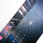 Nidecker Venus Plus Women's Snowboard 2025 - A black snowboard with a pink and blue abstract design, featuring a woman riding it in the background.