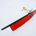 Nidecker Sensor Women's Snowboard 2025 - A snowboard with a black base and white stripes on the top, featuring a rider in a yellow jacket riding it down a snowy slope.