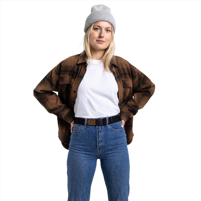 A woman wearing a black and brown checkered shirt over a white t-shirt and blue jeans, showing a black belt with a brown patch around her waist. She stands with her hands on her hips, looking at the camera.