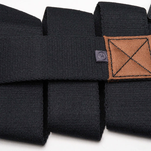 A close-up of a black fabric belt with a brown rectangular patch featuring stitched detailing. The belt is folded multiple times, highlighting its texture and the small logo tag next to the patch.