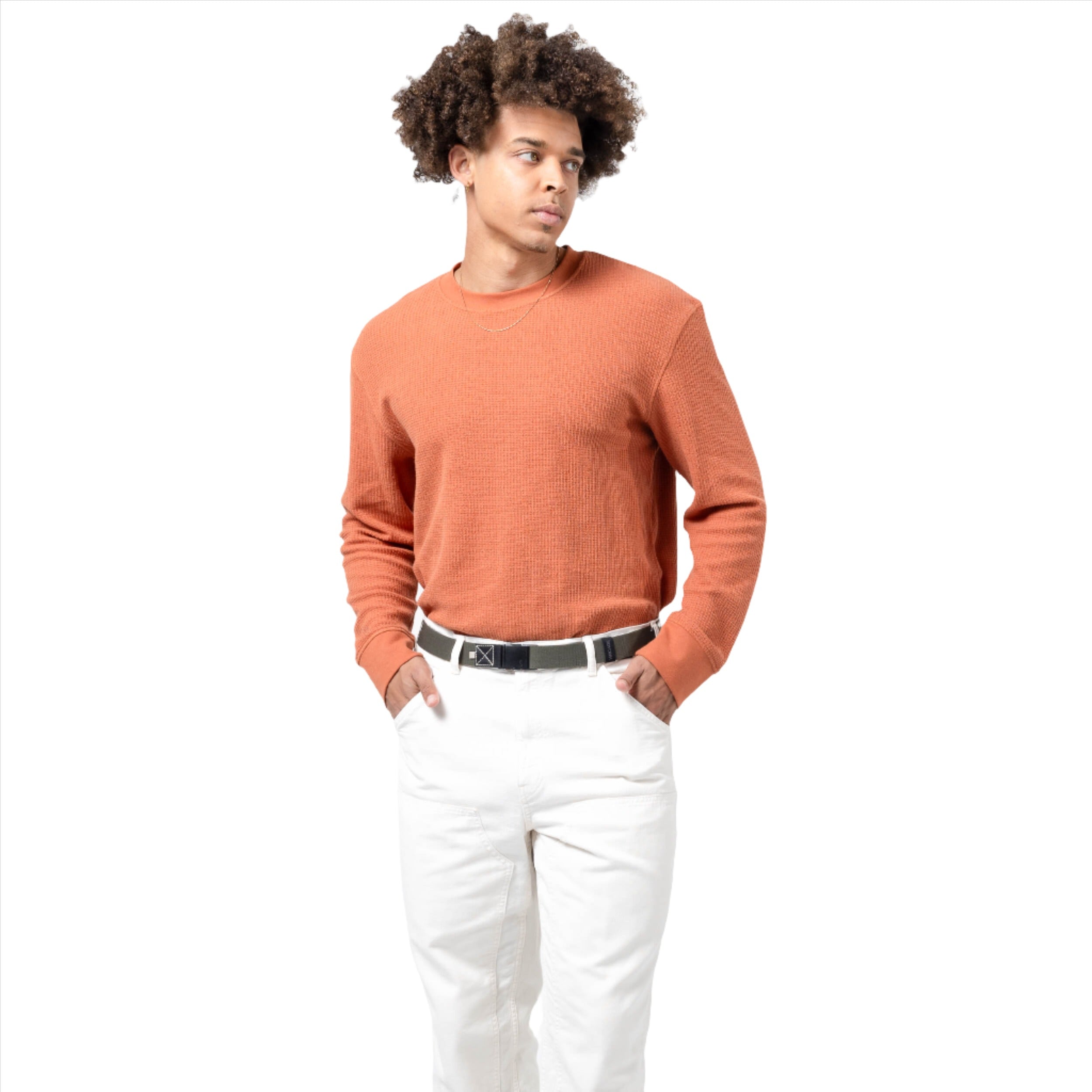 A man wearing an orange sweater and white pants, showing an ivy green belt with a black patch and plastic buckle around his waist. He stands with his hands in his pockets, looking to the side.