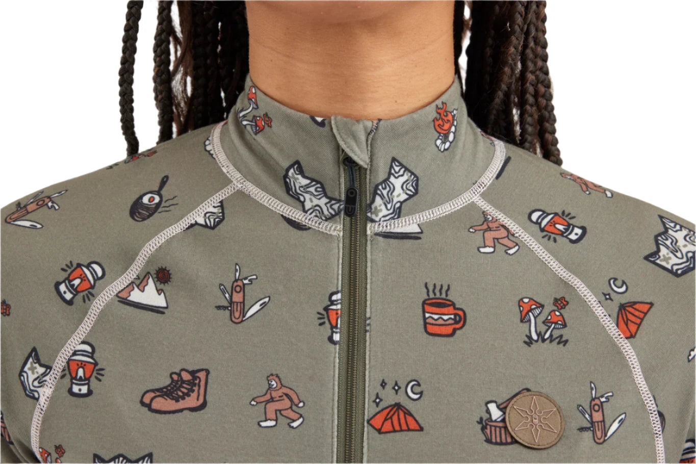 Airblaster Women's Hoodless Ninja Suit 2025 - A sage green onesie with a repeating pattern of cartoon-style illustrations, including skiers, snowboarders, and various winter-themed icons.