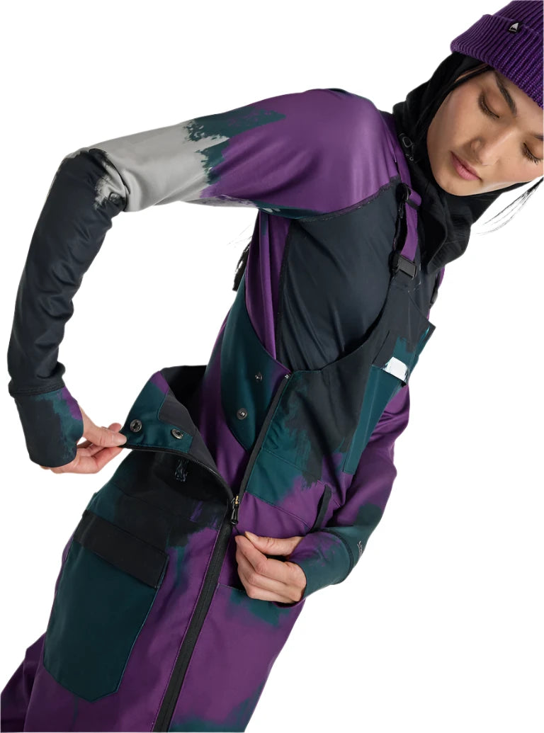 Burton Women's Avalon Stretch 2L Bib Pant 2025 - A purple and black bib pant with a snowflake pattern on the left leg, worn by a model posing in front of a snowy background.