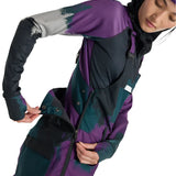 Burton Women's Avalon Stretch 2L Bib Pant 2025 - A purple and black bib pant with a snowflake pattern on the left leg, worn by a model posing in front of a snowy background.