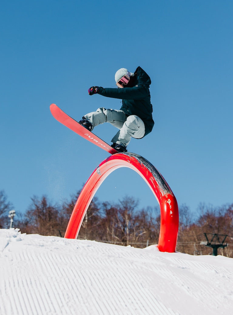 Burton Women's Pillowline GORE-TEX Insulated Jacket 2025 - A woman in a black jacket and gray snow pants is riding a snowboard on top of an orange archway, with trees in the background.