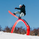 Burton Women's Pillowline GORE-TEX Insulated Jacket 2025 - A woman in a black jacket and gray snow pants is riding a snowboard on top of an orange archway, with trees in the background.