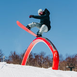 Burton Women's Pillowline GORE-TEX Insulated Jacket 2025 - A woman in a black jacket and gray snow pants is riding a snowboard on top of an orange archway, with trees in the background.