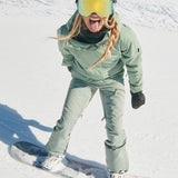 Roxy Women's Illumine Warmlink Jacket - A sage green jacket with a black collar and two zippered pockets on the front, worn by a model posing in front of a snowy background.