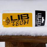 Lib Tech Wax Scrapper - A yellow plastic scraper with a hole at one end, featuring black lettering and a simple stick figure drawing of a person wearing a pointed hat.