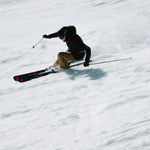 Völkl Peregrine 82 Skis with Lowride 13FR Bindings - A skier in a black jacket and brown pants is skiing down the mountain on a pair of black skis with red accents.