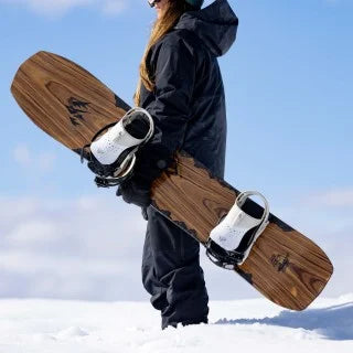 Jones Women's Flagship Snowboard 2024 - A brown snowboard with a black logo on top, held by a woman in a dark blue jacket and pants.