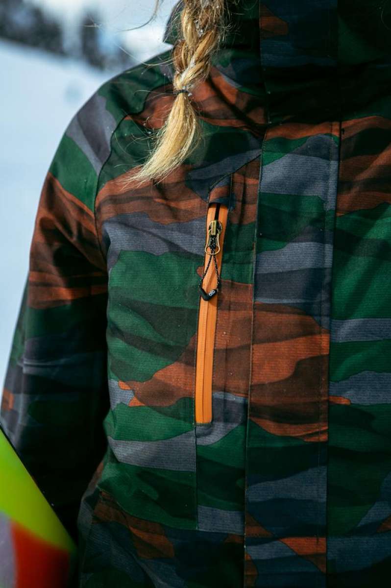 The 686 Willow Insulated Jacket, a camouflage jacket with an orange zipper and accents, is worn by a woman in front view.
