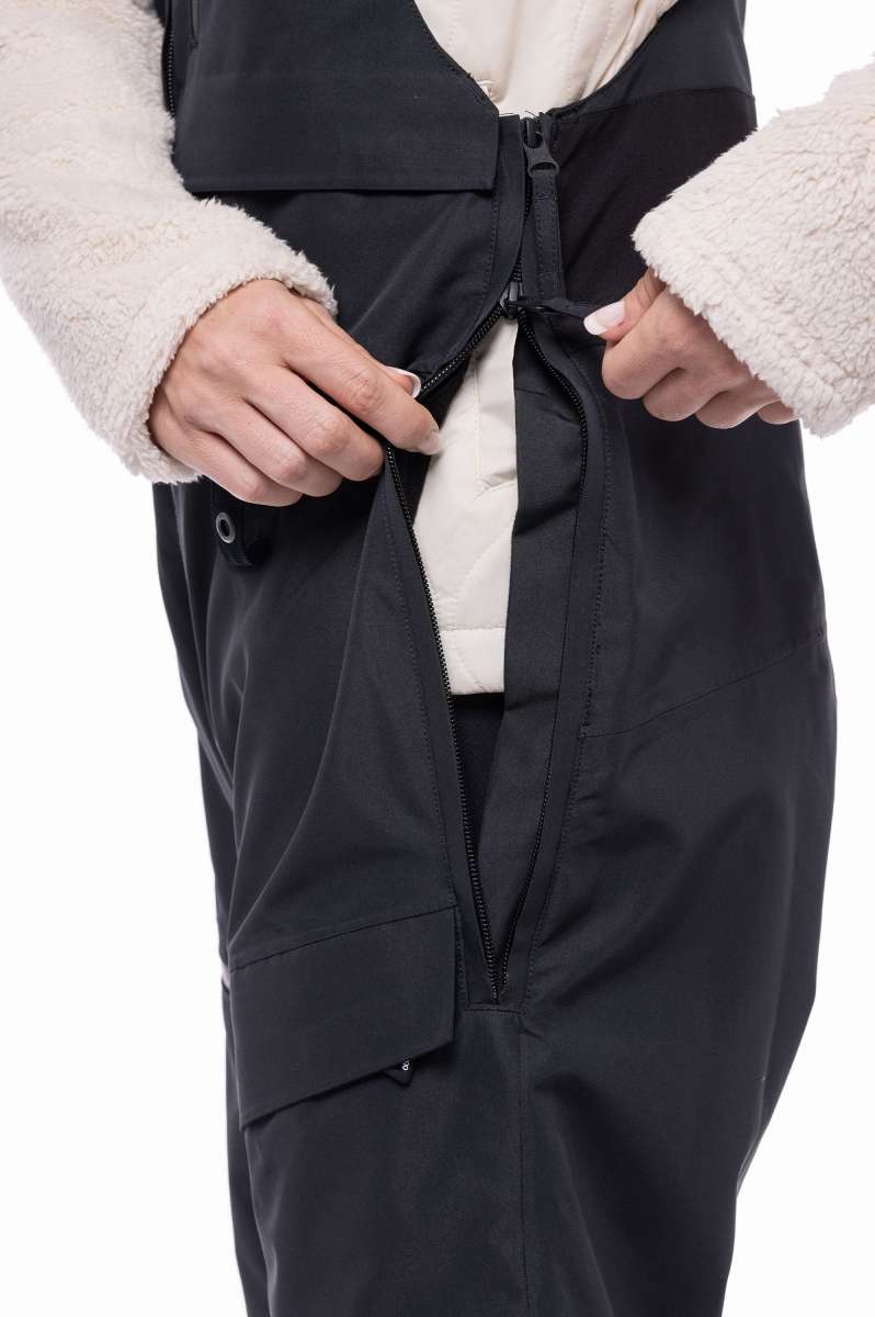 The 686 Women's Geode Thermagraph Bib 2023 has a black bib with an insulated cream-colored hood and white faux fur trim.
