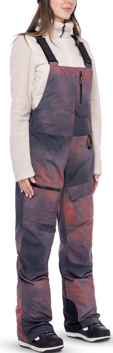 The women's bib snow pants are dark gray with a pinkish-red overlay and have zippered front pockets.