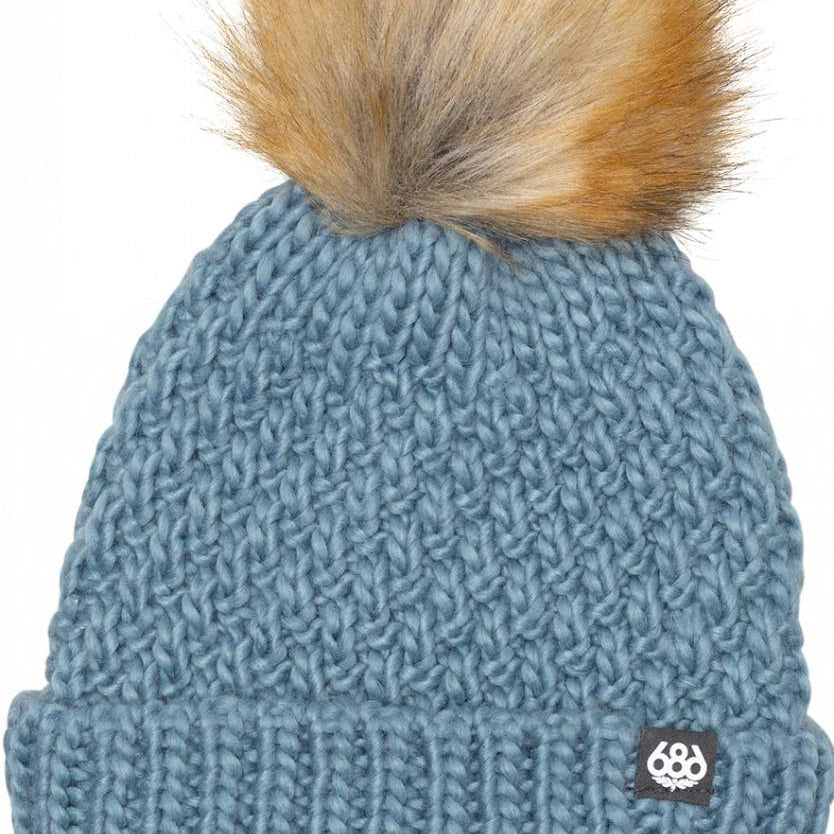 The 686 Ladies Majesty Cable Knit Beanie 2024 is light blue with a ribbed cuff and a large brown faux fur pom-pom on top.