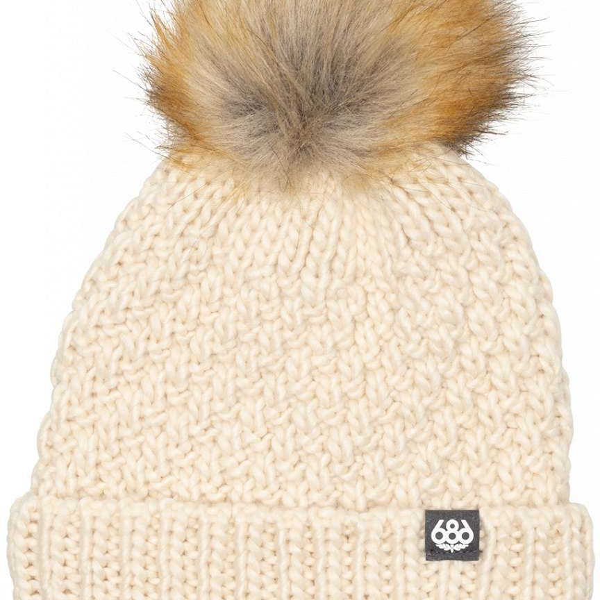 The 686 Majesty Beanie is crafted from cream-colored cable knit fabric and features a fluffy brown faux fur pom-pom on top.