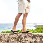 Reef Men's Fanning Sandal - A man wearing khaki shorts is standing on a rocky surface overlooking the ocean, facing away from the camera and showing only his legs and feet. He is wearing black sandals with white logos on the straps.