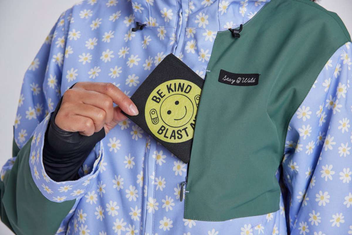 Airblaster Women's Freedom Insulated Suit 2024 - A woman wearing a light blue snowsuit with white daisies and yellow centers, holding it up to show off the front zipper and black logo on the chest.