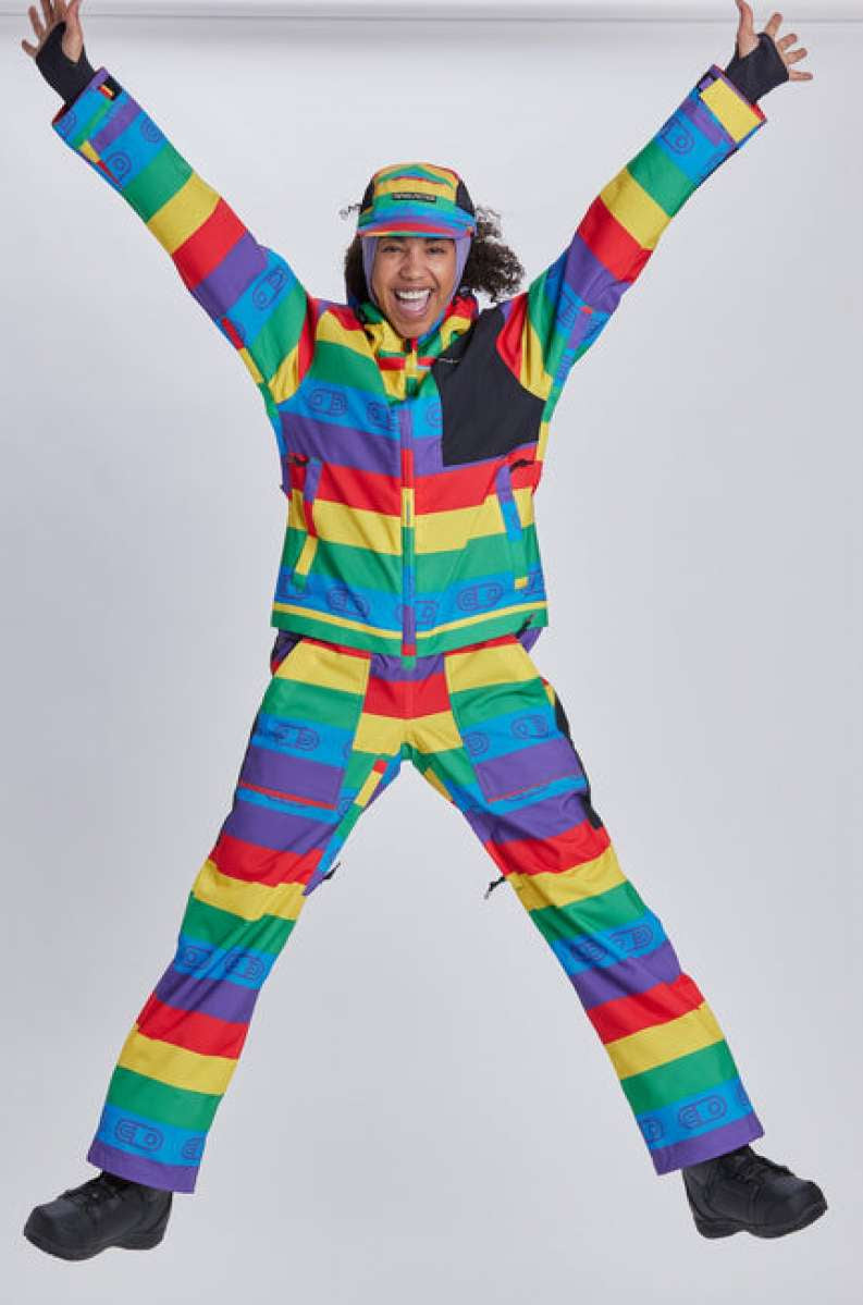 Airblaster Women's Freedom Stretch Suit 2024 - A woman wearing a rainbow-striped, multicolored snowsuit with black accents and a matching hat.