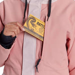 Airblaster Women's Stay Wild Insulated Parka - A woman wearing a dusty rose parka with white trim and black drawstrings, holding a yellow card in her hand.
