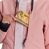 Airblaster Women's Stay Wild Insulated Parka - A woman wearing a dusty rose parka with white trim and black drawstrings, holding a yellow card in her hand.