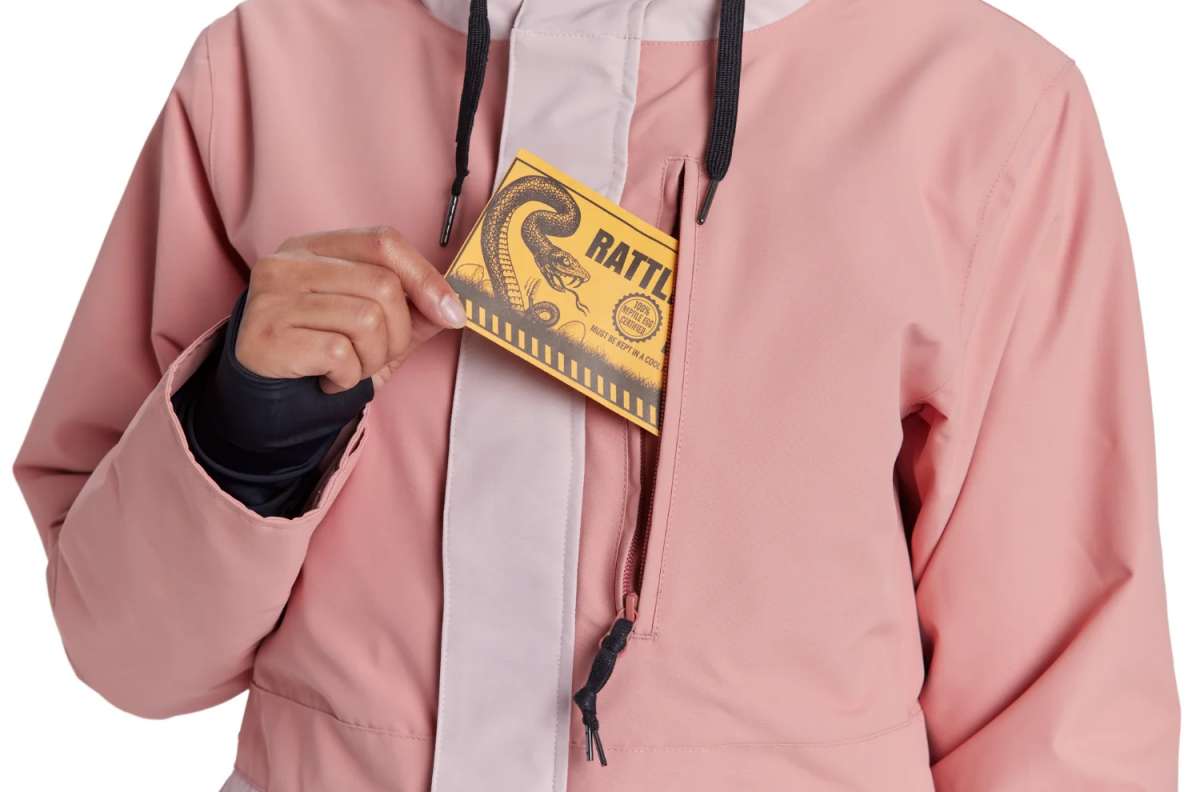 Airblaster Women's Stay Wild Insulated Parka - A woman wearing a dusty rose parka with white trim and black drawstrings, holding a yellow card in her hand.