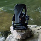 Arbor Cypress Snowboard Bindings 2024 - A pair of black bindings with a distinctive orange accent on the highback, sitting atop a rock near a body of water.