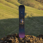 Arbor Draft Camber Snowboard 2024 - A black snowboard with a white logo and blue accents on top of a rocky surface.
