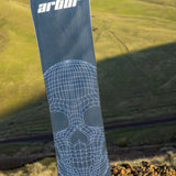Arbor Draft Camber Snowboard 2024 - A black snowboard with a white geometric pattern on top of a rocky hillside.