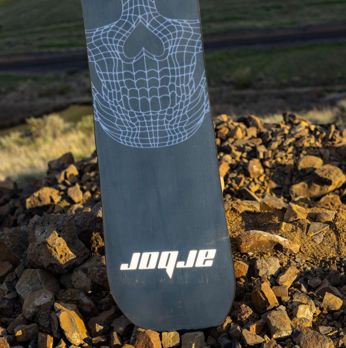 Arbor Draft Camber Snowboard 2024 - A black snowboard with a white spiderweb design on top of a pile of rocks.