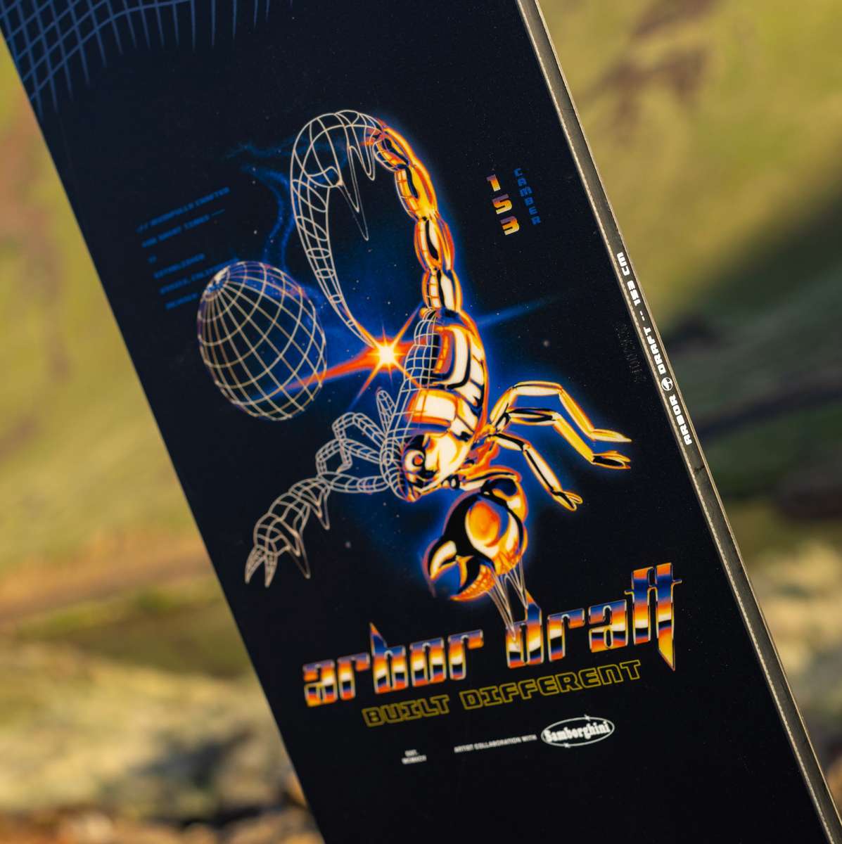 Arbor Draft Camber Snowboard 2024 - A black snowboard with a white and blue geometric pattern on top of a blurred background.