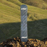 Arbor Draft Camber Snowboard 2024 - A black snowboard with a white grid pattern on top of a rocky hillside.