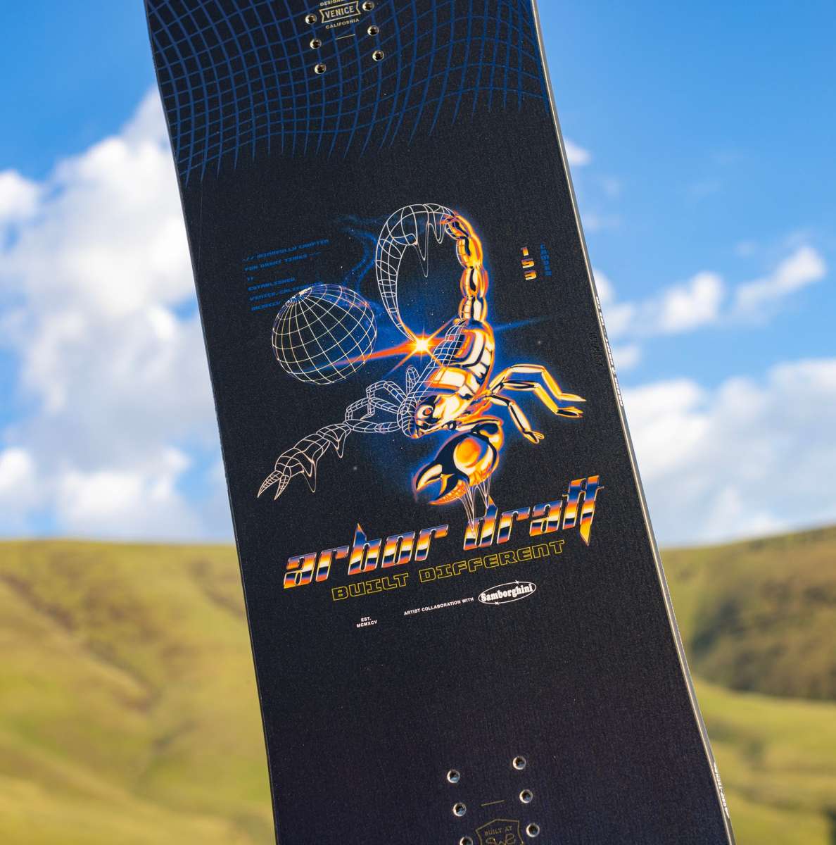 Arbor Draft Camber Snowboard 2024 - A black snowboard with a white and blue geometric pattern on top of a snowy mountain.