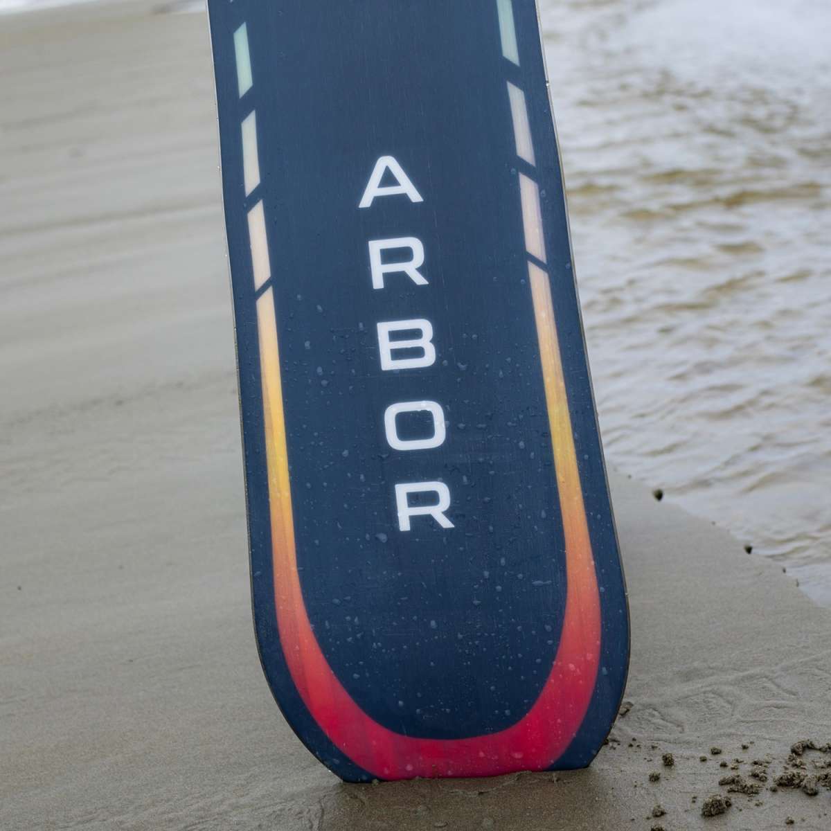 Arbor Formula Rocker Snowboard 2024 - A black snowboard with a red and yellow stripe along its bottom edge, standing upright on a sandy surface.