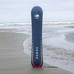 Arbor Formula Rocker Snowboard 2024 - A dark blue snowboard with a white logo featuring an arched tree, standing upright on a sandy beach with gentle waves rolling in behind it.