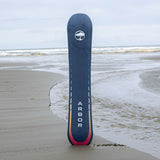 Arbor Formula Rocker Snowboard 2024 - A dark blue snowboard with a white logo featuring an arched tree, standing upright on a sandy beach with gentle waves rolling in behind it.
