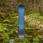 Arbor Foundation Rocker Snowboard 2024 - A blue snowboard with a white logo at the top and a black logo near the bottom, standing on rocks in front of a mossy stream.