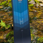 Arbor Foundation Rocker Snowboard 2024 - A blue snowboard with a geometric pattern on top and a black bottom, standing in a stream of water.