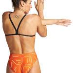 Arena Women's Puzzled Challenge Back One Piece Swimsuit - A woman with brown hair in a bun, wearing an orange one-piece swimsuit with a geometric pattern of white lines and shapes on the fabric, facing away from the camera.
