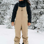 Burton Women's AK Gore-Tex Kimmy Bib Pant 2023 - A pair of tan snow pants with black accents and a zippered pocket on the thigh.