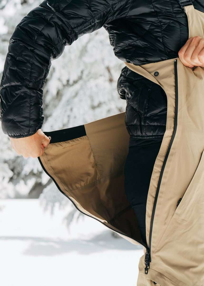 Burton Women's AK Gore-Tex Kimmy Bib Pant 2023 - A black bib pant with a zippered fly and adjustable waistband, featuring a snow-covered pine tree in the background.