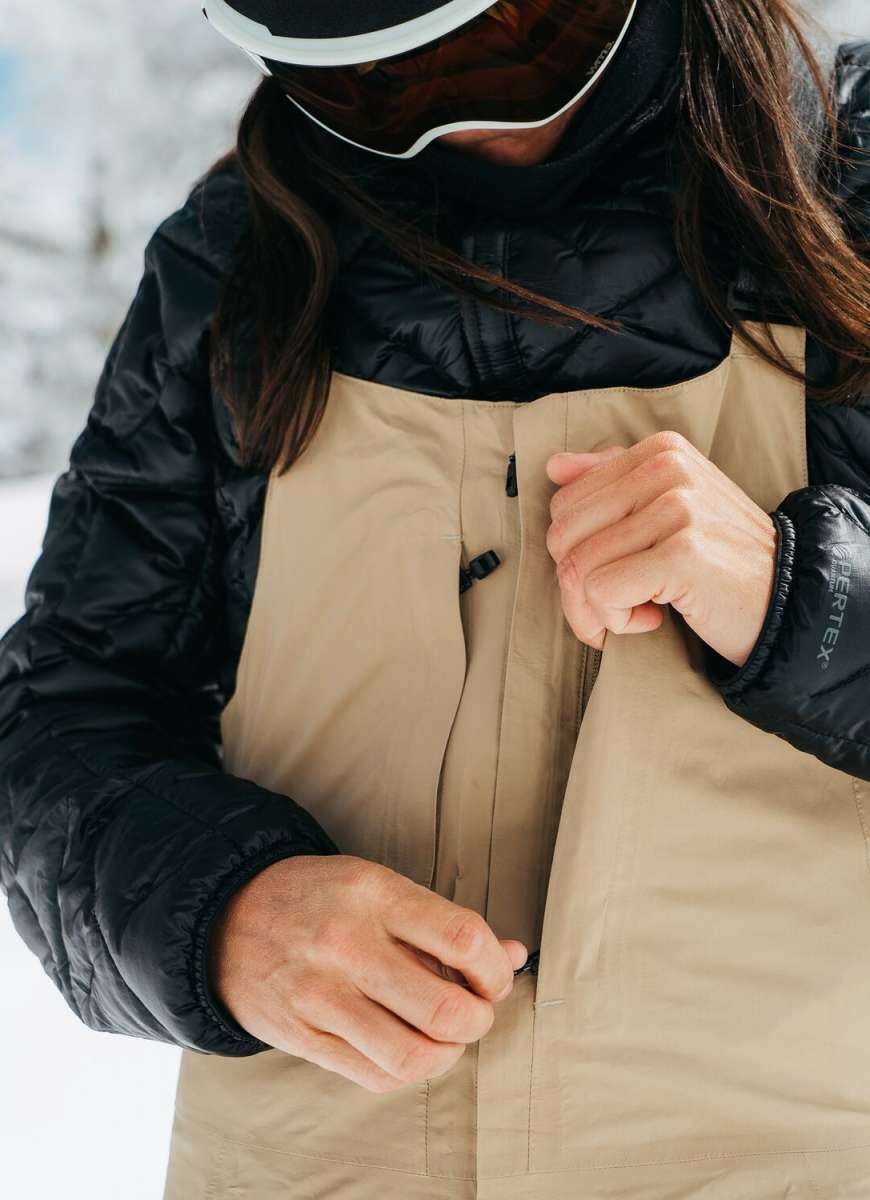 Burton Women's AK Gore-Tex Kimmy Bib Pant 2023 - A tan bib pant with a black zipper and buckle on the side, featuring a zippered leg opening.
