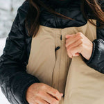 Burton Women's AK Gore-Tex Kimmy Bib Pant 2023 - A tan bib pant with a black zipper and buckle on the side, featuring a zippered leg opening.