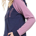 Roxy Women's Liberty Pullover Technical Hoodie 2020 - A woman with long blonde hair wearing a gray knit beanie and a navy-blue hoodie with pink sleeves, posing slightly to her left.