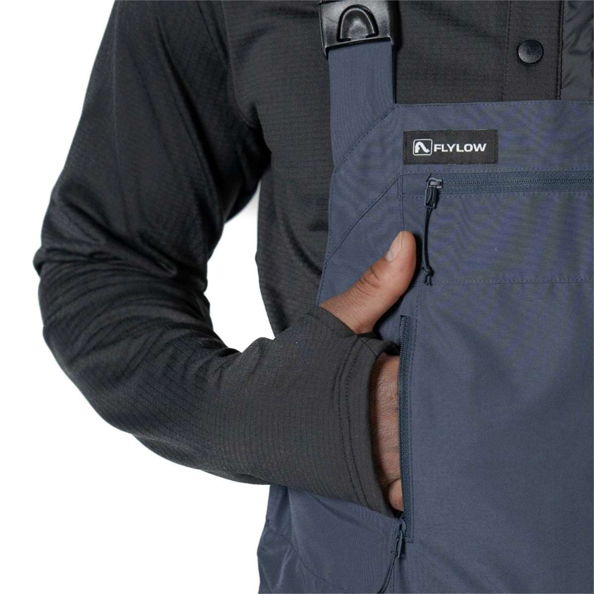 Flylow Firebird Shell Bib Pants 2024 - A pair of dark gray bib pants with a black logo patch on the left thigh and a zippered pocket on the right side.