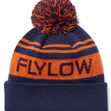 Flylow OG Pom Beanie 2024 - A navy blue beanie with an orange stripe and a large pom-pom on top.